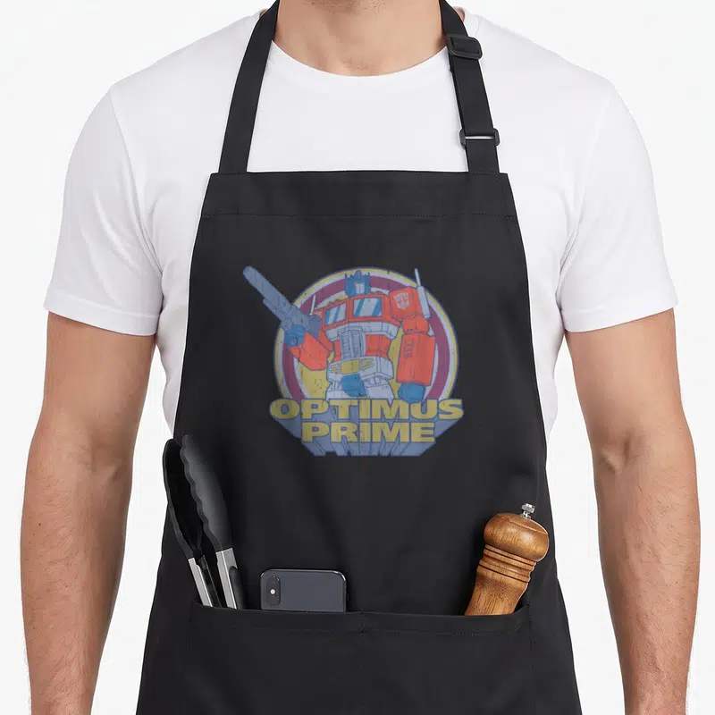 Cute portrait transformer apron - Optimus Prime robot in red blue yellow with 3D text and concentric circles — shown on a black cotton canvas apron against a white background