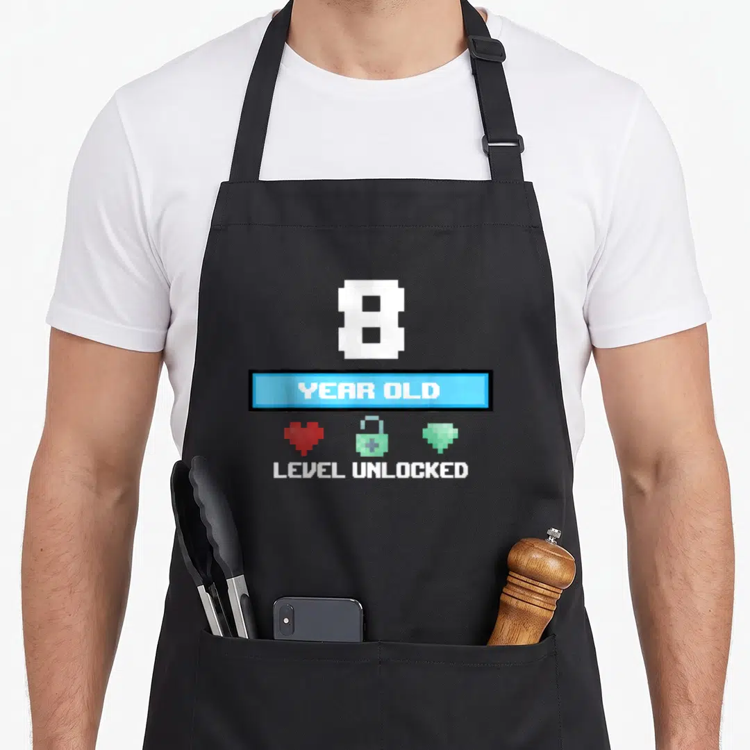8th level birthday apron - Pixel art number 8, year old banner, heart lock diamond icons, level unlocked text — shown on a black cotton canvas apron against a white background
