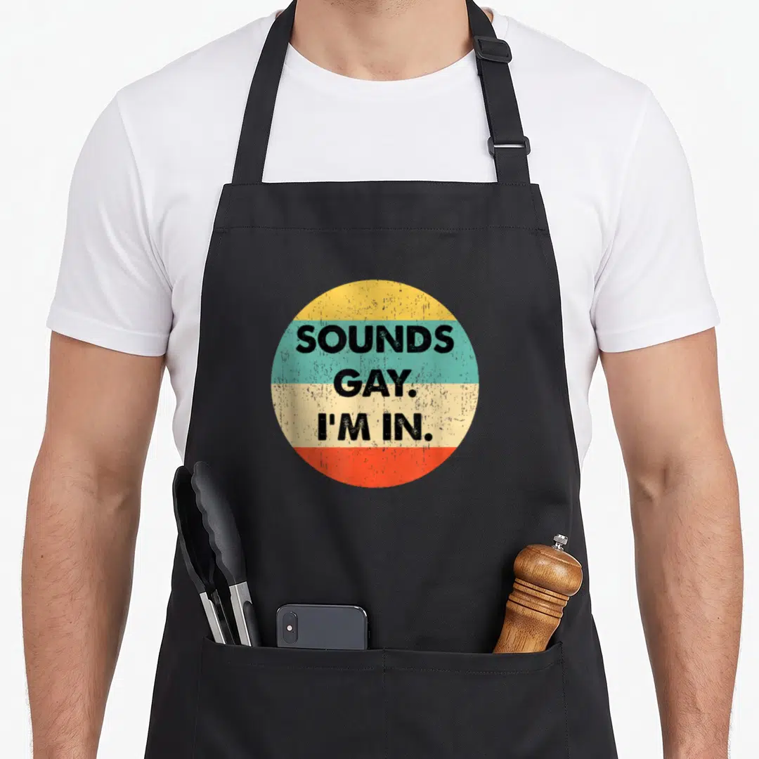 Funny sound gay apron - SOUNDS GAY. I'M IN. text in circle with yellow teal beige red stripes — shown on a black cotton canvas apron against a white background