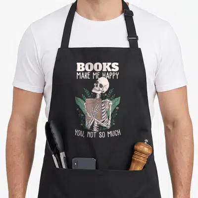 Book skeleton apron - Skeleton holding brown book with green leaves and pink flowers, beige text — shown on a black cotton canvas apron against a white background
