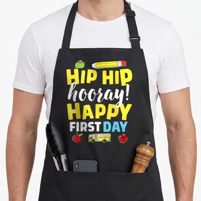 School teacher apron for men - Stacked text HIP HIP hooray HAPPY FIRST DAY with lunchbox pencil apple bus and book — shown on a black cotton canvas apron against a white background
