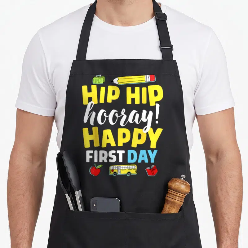 School teacher apron for men - Stacked text HIP HIP hooray HAPPY FIRST DAY with lunchbox pencil apple bus and book — shown on a black cotton canvas apron against a white background
