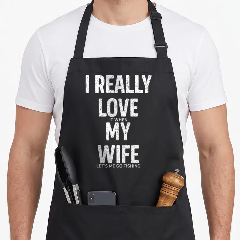 Wife joke fishing aprons - Stacked white text reading I really love it when my wife lets me go fishing — shown on a black cotton canvas apron against a white background