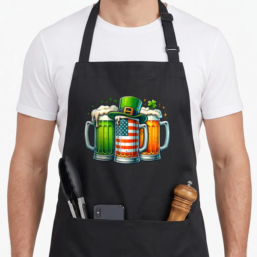 Drinking ireland apron - Three beer mugs with leprechaun hat and shamrocks in green orange blue red white cartoon style — shown on a black cotton canvas apron against a white background