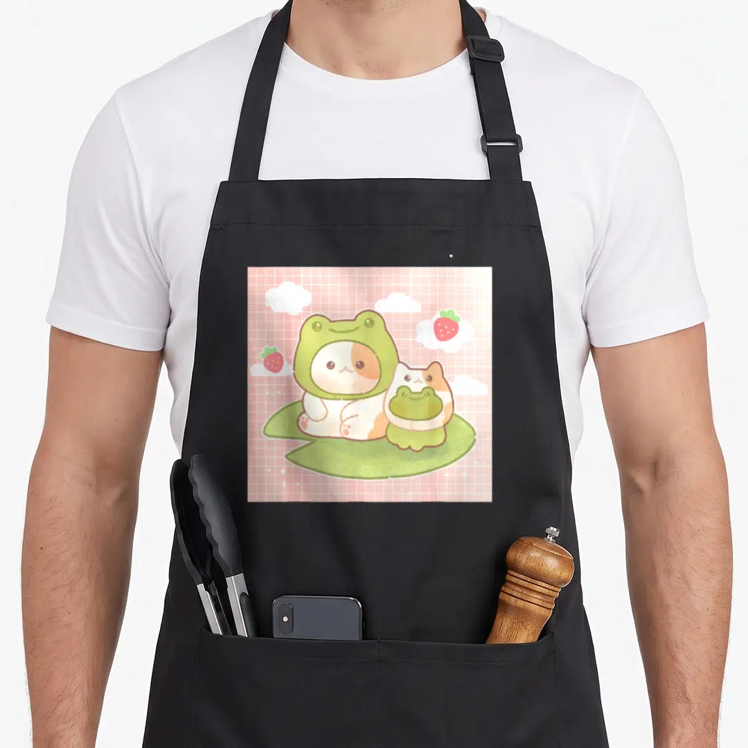 90s cottagecore apron - Two cartoon cats in green frog hats on a lily pad with strawberries on a pink grid — shown on a black cotton canvas apron against a white background