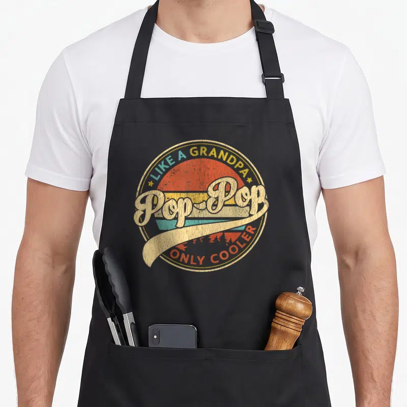 Cooler father day apron - Retro circular graphic with Pop-Pop text over yellow orange teal striped sunset background — shown on a black cotton canvas apron against a white background