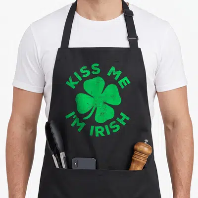 Kiss irish aprons - Green distressed shamrock with arched KISS ME I'M IRISH text on black fabric — shown on a black cotton canvas apron against a white background