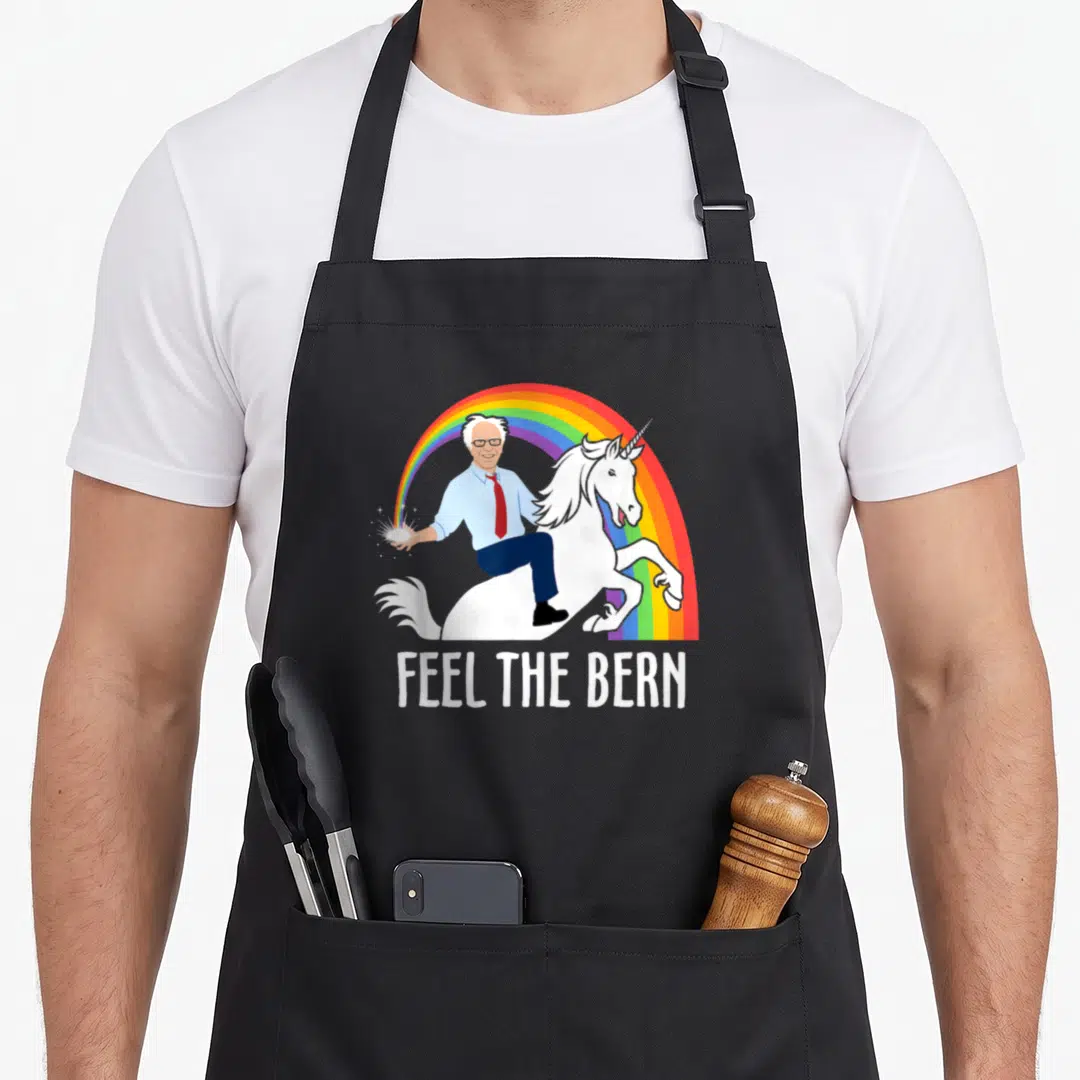 Feel unicorn apron - Man riding white unicorn in front of rainbow with FEEL THE BERN text below — shown on a black cotton canvas apron against a white background
