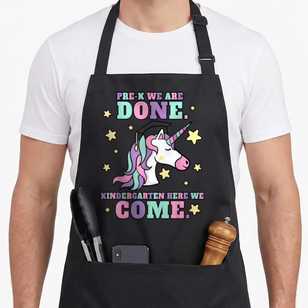 Pre unicorn apron - Cartoon unicorn in graduation cap with PRE-K WE ARE DONE text and stars — shown on a black cotton canvas apron against a white background