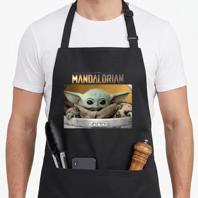 Screenshot pod aprons - Groguppy in white pod below gold The Mandalorian text with tiny figure in letter — shown on a black cotton canvas apron against a white background