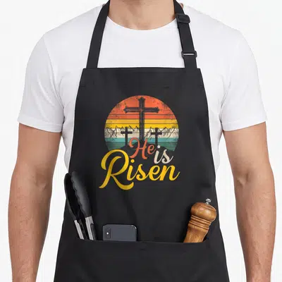 Risen christian easter apron - Three grey crosses on a hill before a striped retro sun with He is Risen script text — shown on a black cotton canvas apron against a white background