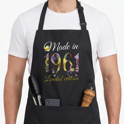 65th vintage floral apron - Watercolor floral year 1961 text with yellow rose and limited edition script — shown on a black cotton canvas apron against a white background