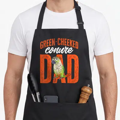 Cheeked bird apron - Cartoon green cheeked conure parrot standing in front of orange DAD text — shown on a black cotton canvas apron against a white background