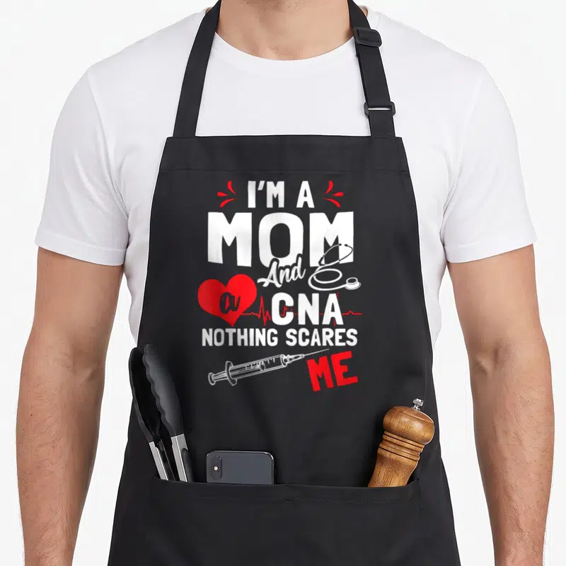 Scare white nurse apron - Stacked I'm a Mom CNA text with red heart EKG line stethoscope and syringe graphics — shown on a black cotton canvas apron against a white background