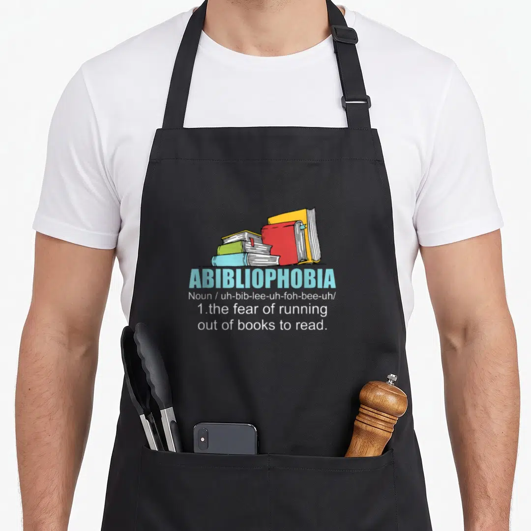 Library nerd apron - Abibliophobia dictionary definition with cartoon stack of blue red and yellow books — shown on a black cotton canvas apron against a white background