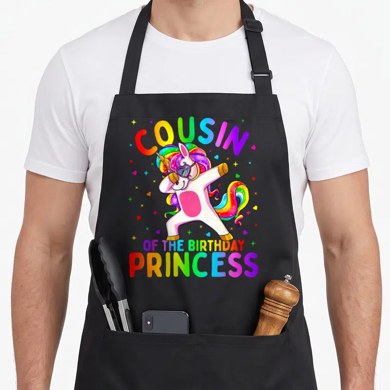 Cousin princess apron - Dabbing unicorn with rainbow mane and heart sunglasses surrounded by stars and hearts — shown on a black cotton canvas apron against a white background