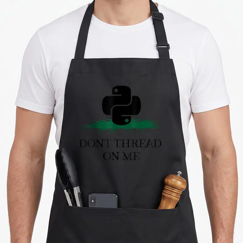 Thread python jokes apron - Black python logo on green grass above DONT THREAD ON ME text — shown on a black cotton canvas apron against a white background