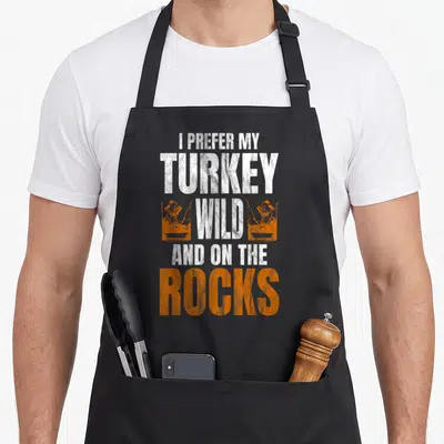 Prefer turkey apron - Stacked white and orange text reading I Prefer My Turkey Wild And On The Rocks flanked by two — shown on a black cotton canvas apron against a white background