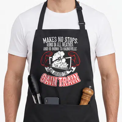 Funny train gains apron - White steam train flanked by red muscular arms with gain pun text — shown on a black cotton canvas apron against a white background