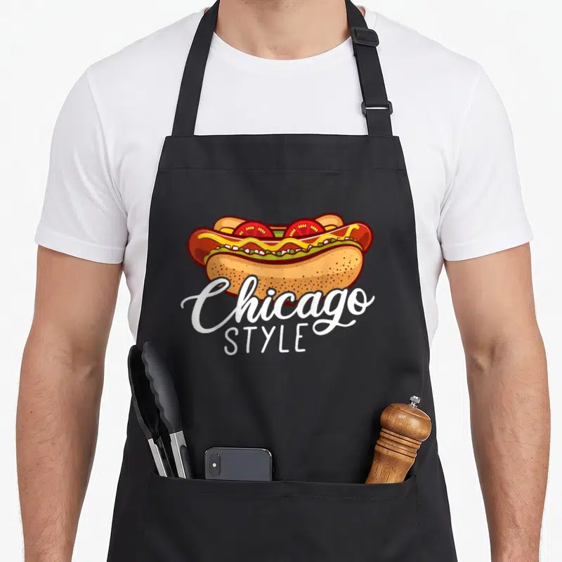 Chicago hotdog hot dog apron - Cartoon hot dog with mustard tomato relish and Chicago STYLE text below — shown on a black cotton canvas apron against a white background