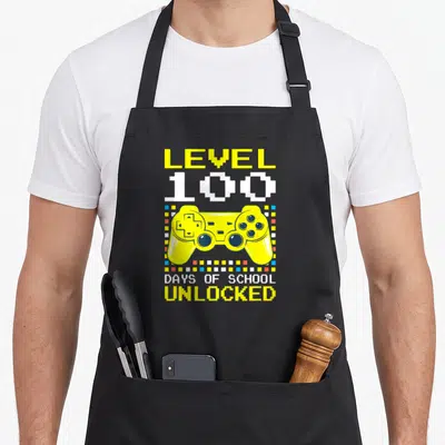 Level school video game apron - Yellow pixelated game controller with LEVEL 100 DAYS OF SCHOOL UNLOCKED text — shown on a black cotton canvas apron against a white background