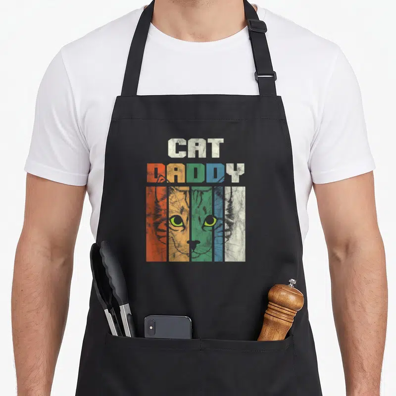 Pet cat dad apron - Retro cat face with stacked CAT DADDY text in orange yellow green blue and white vertical stripes — shown on a black cotton canvas apron against a white background