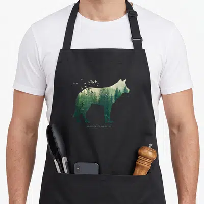 Protect preserves apron - Wolf silhouette filled with green forest trees and white flying birds above text — shown on a black cotton canvas apron against a white background