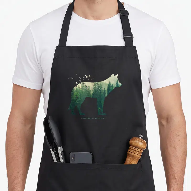 Protect preserves apron - Wolf silhouette filled with green forest trees and white flying birds above text — shown on a black cotton canvas apron against a white background