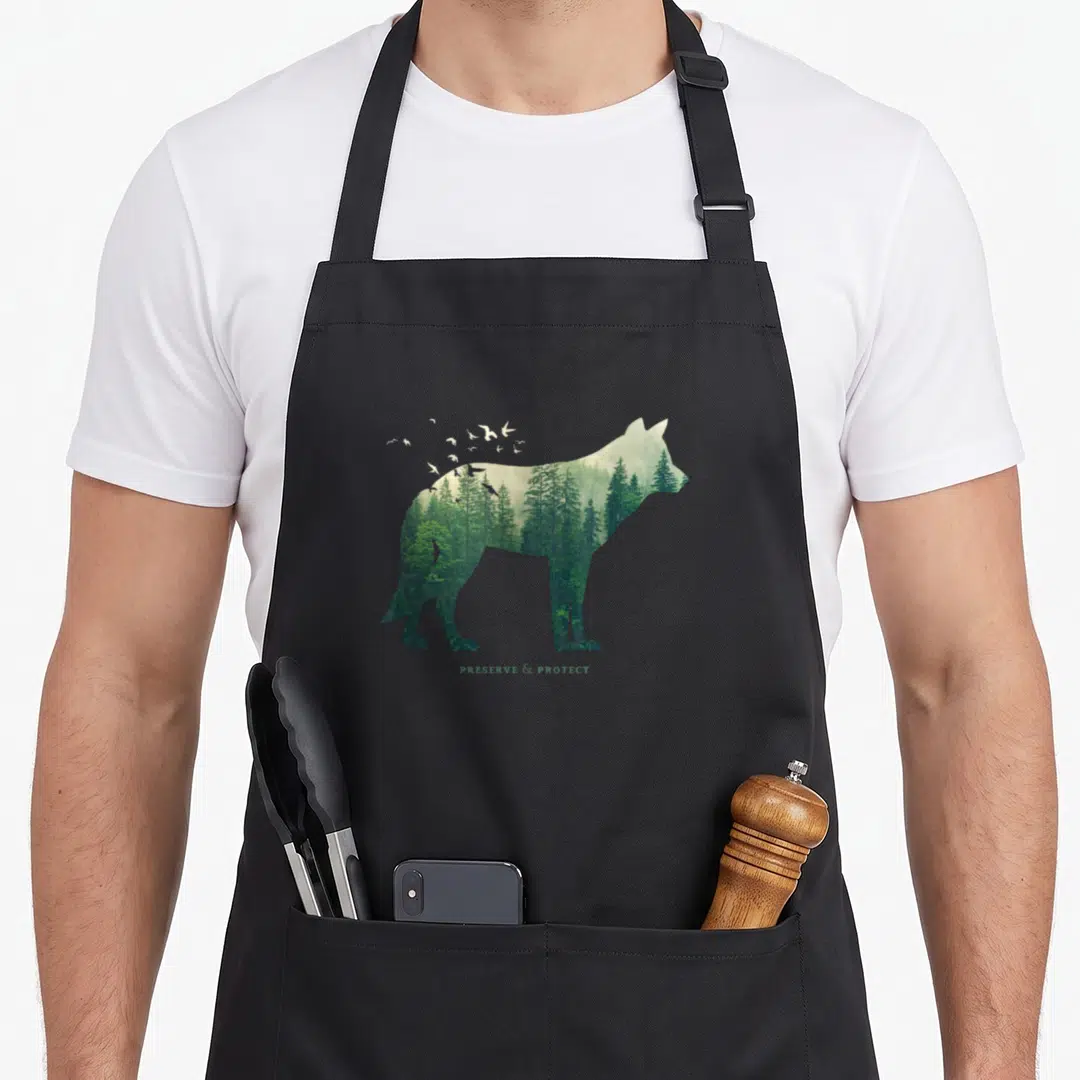 Protect preserves apron - Wolf silhouette filled with green forest trees and white flying birds above text — shown on a black cotton canvas apron against a white background
