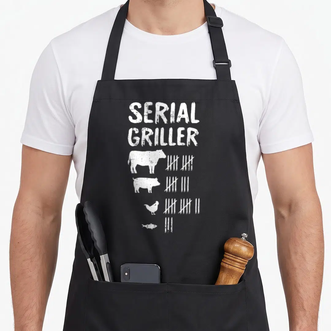 Serial master griller apron - White distressed text Serial Griller above four animals with tally marks on black fabric — shown on a black cotton canvas apron against a white background