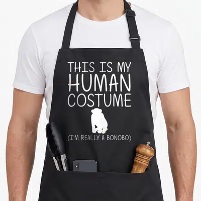 Bonobo halloween apron - White text reading THIS IS MY HUMAN COSTUME above a bonobo silhouette — shown on a black cotton canvas apron against a white background