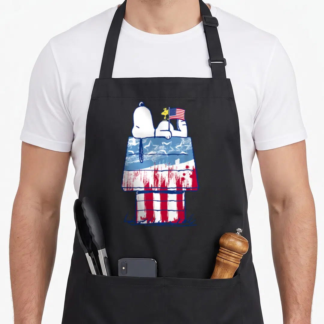 Funny wood apron - Snoopy and Woodstock lying on a doghouse painted with red, white, and blue stars and stripes — shown on a black cotton canvas apron against a white background