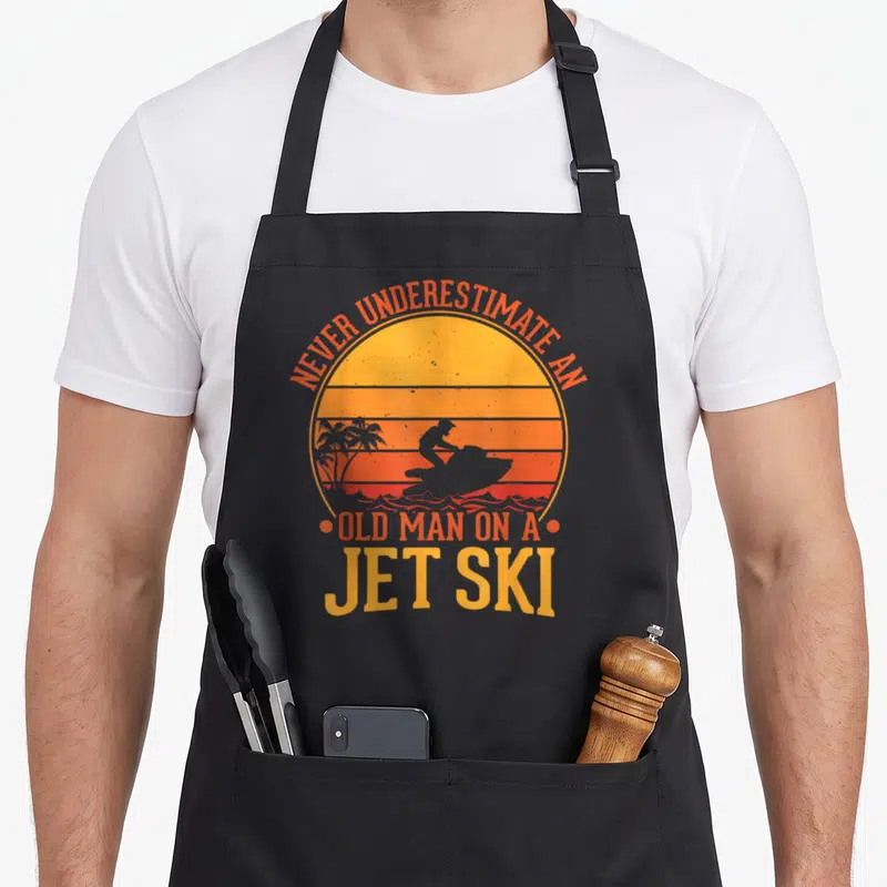 Water jet ski apron - Retro sunset circle with jet ski silhouette and orange text reading Never Underestimate An Old — shown on a black cotton canvas apron against a white background