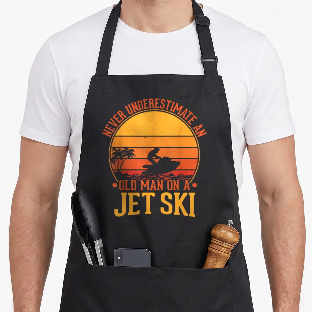 Water jet ski apron - Retro sunset circle with jet ski silhouette and orange text reading Never Underestimate An Old — shown on a black cotton canvas apron against a white background