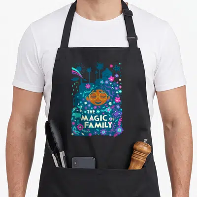 Family magic apron - Stylized girl face with hair blending into city and flowers, stacked text reading THE MAGIC — shown on a black cotton canvas apron against a white background