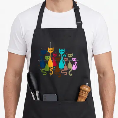 Funny kitten cat apron - Stylized cats in blue, orange, cyan, and lime green arranged on a grey grid with stars — shown on a black cotton canvas apron against a white background