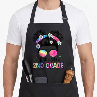 School second 2nd apron - Cartoon girl with tie-dye sunglasses and buns above little Miss 2ND GRADE text with stars — shown on a black cotton canvas apron against a white background