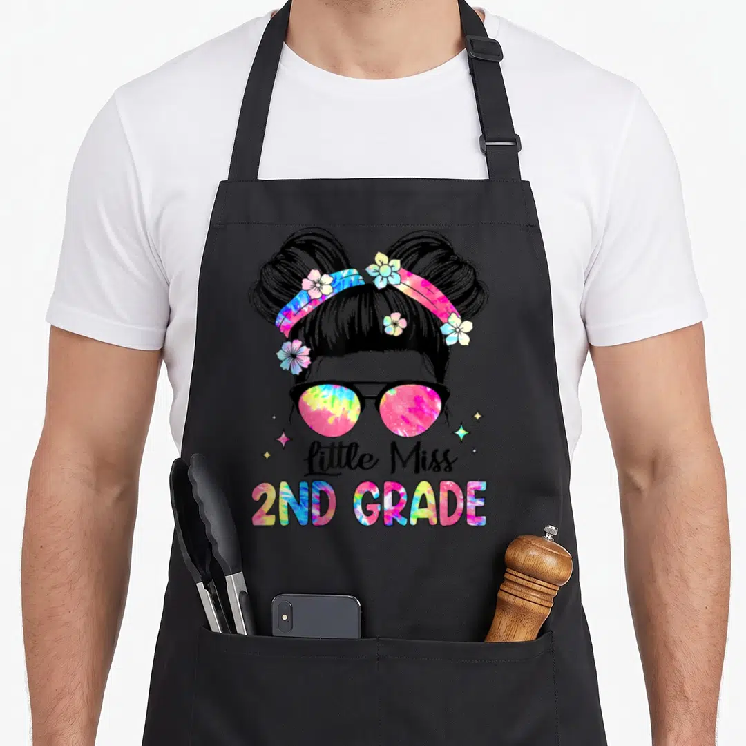School second 2nd apron - Cartoon girl with tie-dye sunglasses and buns above little Miss 2ND GRADE text with stars — shown on a black cotton canvas apron against a white background