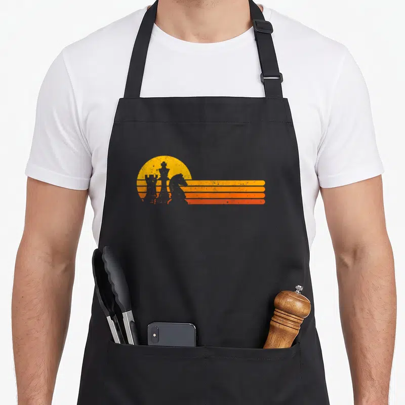 Sunset chess player apron - Retro King, Knight, and Rook silhouettes against yellow sun and orange stripes — shown on a black cotton canvas apron against a white background