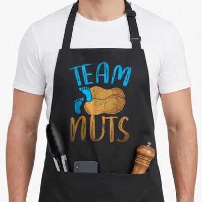 Baby nut gender reveal apron - Two peanuts and footprints with TEAM NUTS text in blue and orange — shown on a black cotton canvas apron against a white background