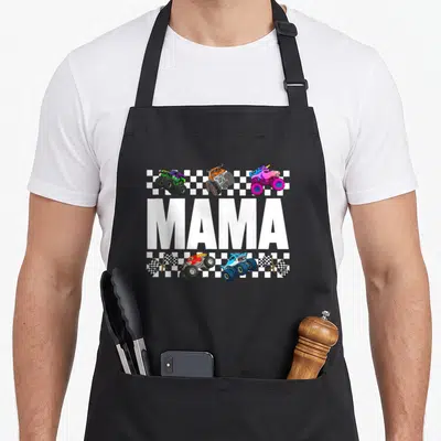Mama crew aprons - MAMA text with five monster trucks over checkered strips and crossed flags — shown on a black cotton canvas apron against a white background