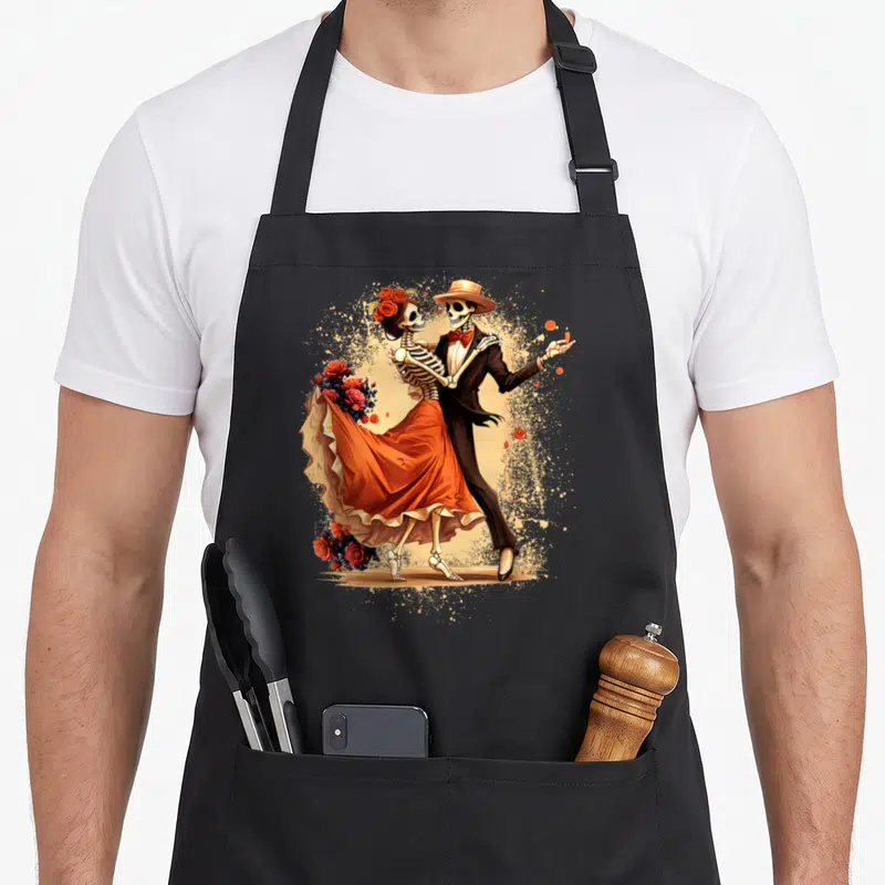 Skeleton day of the dead apron - Cartoon skeleton couple dancing with red roses and tan paint splatters — shown on a black cotton canvas apron against a white background