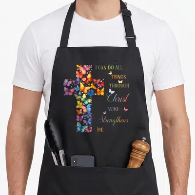 Butterfly cross apron - Cross made of colorful butterflies with stacked Bible verse text in watercolor style — shown on a black cotton canvas apron against a white background