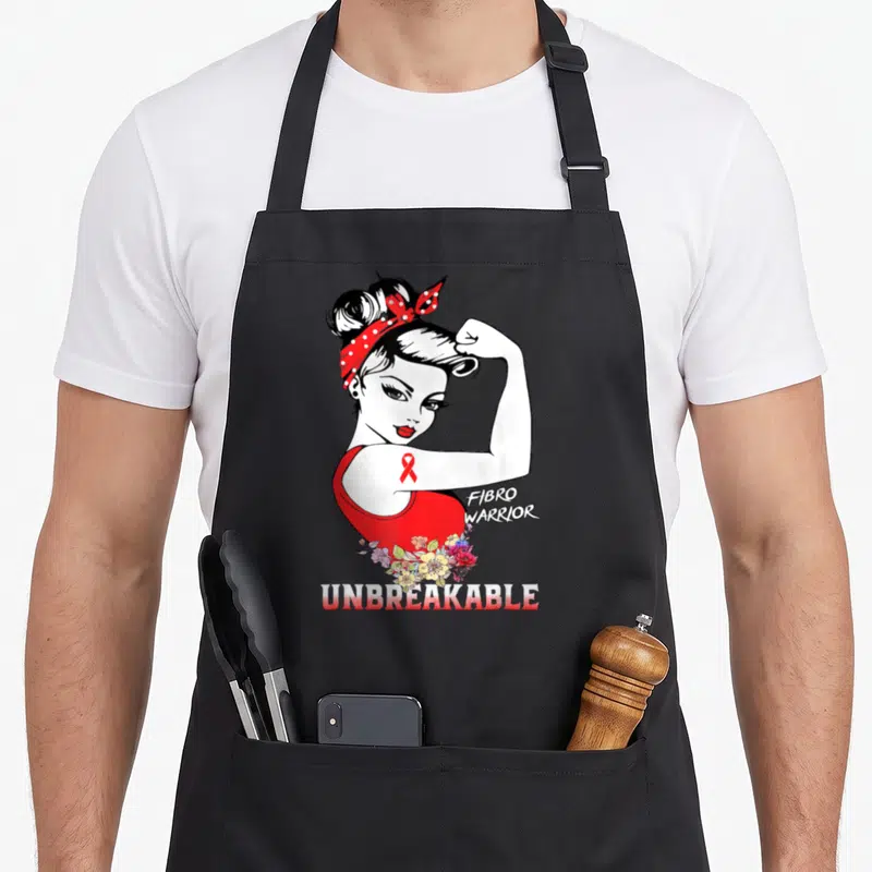 Fibro fibromyalgia awareness apron - Cartoon woman flexing arm with red ribbon, FIBRO WARRIOR text, and UNBREAKABLE — shown on a black cotton canvas apron against a white background