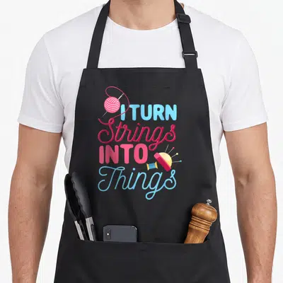 Turn knitting apron - Pink and blue text I Turn Strings Into Things with yarn ball, needle, spool and pincushion — shown on a black cotton canvas apron against a white background