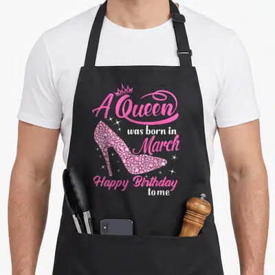 Queen march birthday apron - Pink high heel shoe with crown and A Queen was born in March text — shown on a black cotton canvas apron against a white background