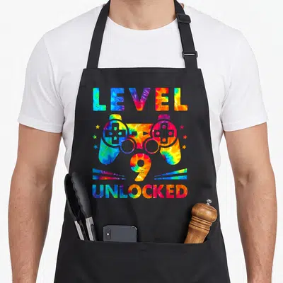 9th tie dye apron - Tie-dye game controller with LEVEL 9 UNLOCKED text in blue red yellow green — shown on a black cotton canvas apron against a white background