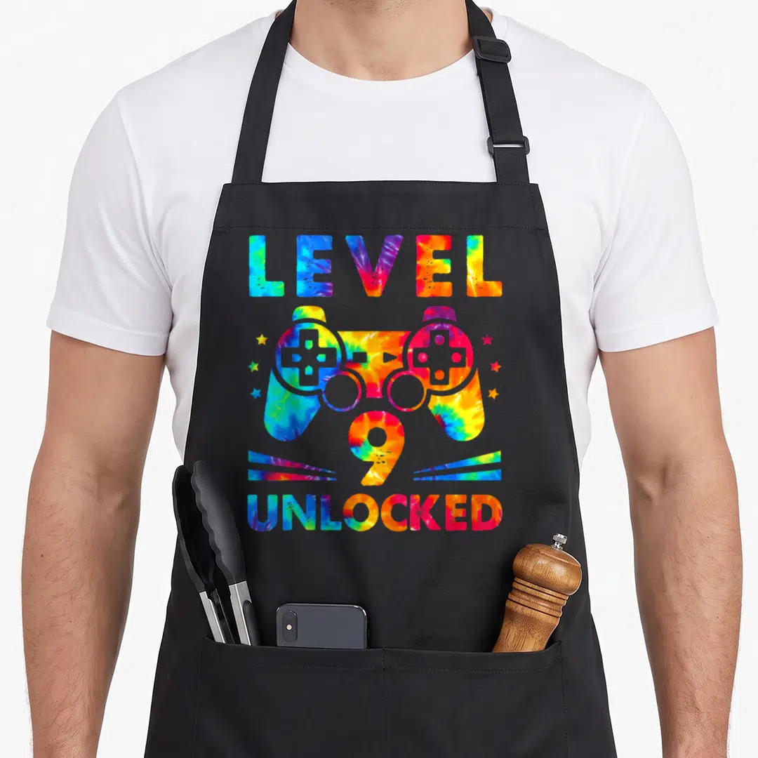 9th tie dye apron - Tie-dye game controller with LEVEL 9 UNLOCKED text in blue red yellow green — shown on a black cotton canvas apron against a white background