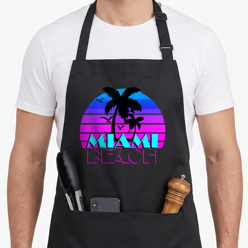 Miami souvenir apron - Retro sun with palm trees and stacked cyan pink MIAMI BEACH text — shown on a black cotton canvas apron against a white background