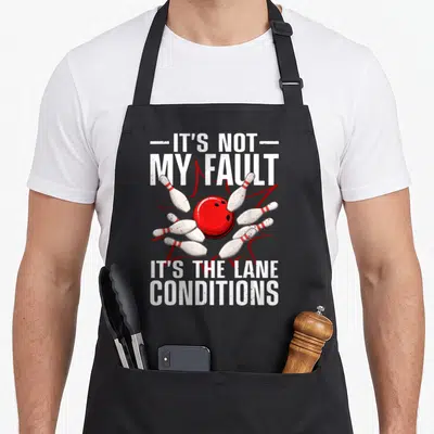 Bowling spare apron - Red bowling ball and scattering pins with It's Not My Fault lane conditions text — shown on a black cotton canvas apron against a white background